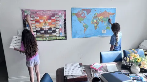 Two kids at a dining room wall, one placing a sticker on a World Cup bracket poster, the other finding a country on a world map